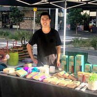 Young man with dark hair wearing a black tshirt standing behind a table filled with products smiling